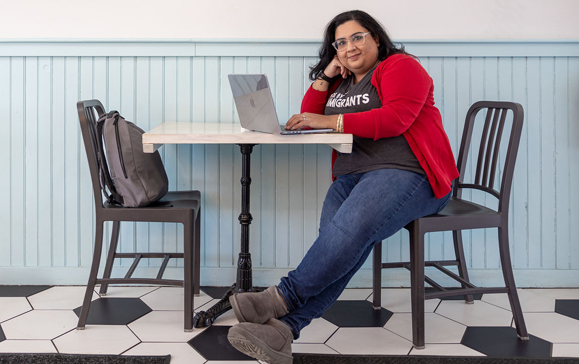 A woman sits at a table in a cafe with her laptop open, and grins at the camera.